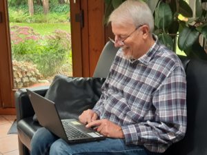 Das Bild zeigt einen lächelnden älteren Mann, der an einem Laptop arbeitet.